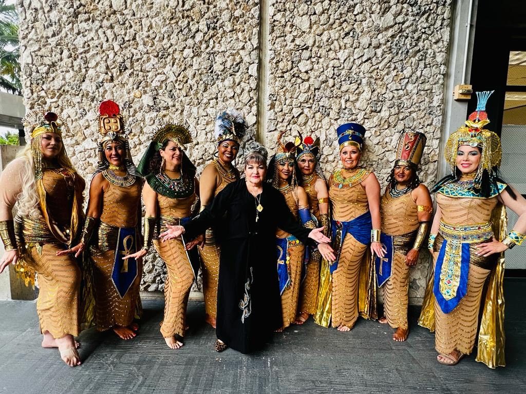 Jihan Jamal in black with arms outstretched, flanked by the ten dancers of Mermaids of the Nile in gold and blue pharaonic costume and headdresses, against a coral stone wall