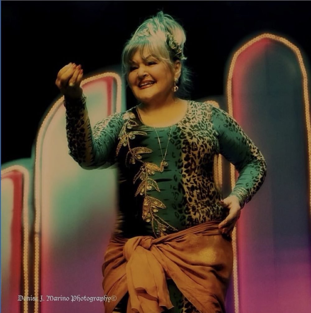 Jihan Jamal on stage, smiling mid-dance in a teal and leopard-print costume with an amber sash